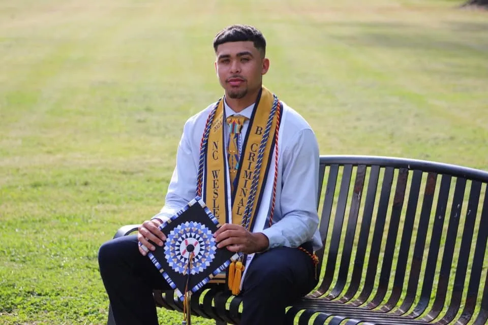 A graduate on a park bench wearing a custom embroidered stole and holding a beaded cap